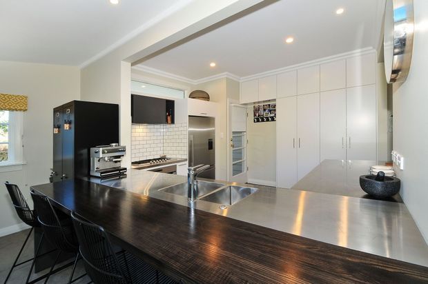 Bright contemporary kitchen with a large dark timber island and integrated seating, stainless-steel benchtops and twin sinks, white cabinetry, white subway-tile splashback, integrated appliances, and recessed ceiling lighting. A dining area is visible at the island, with a partial view toward an adjoining hallway or room.