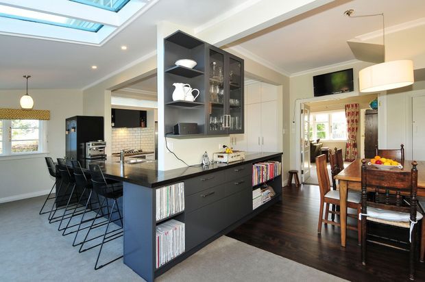 Open-plan residential interior showing a contemporary kitchen, central island with bar seating, and adjacent dining area. The kitchen features dark cabinetry, a black benchtop, white upper cabinetry and white subway-tile backsplash, with skylights providing abundant natural light. A built-in dark media and storage unit separates the kitchen from the dining space, which includes a timber dining table and chairs.