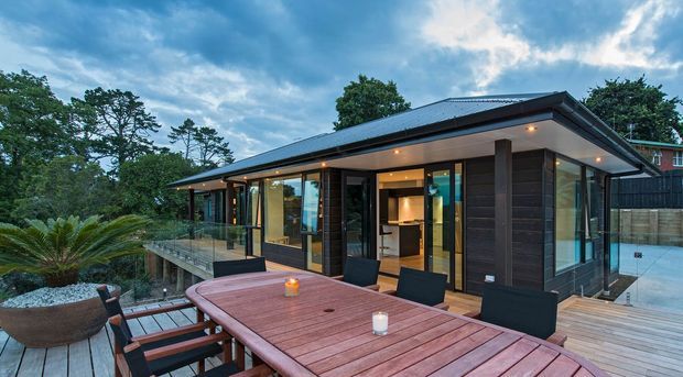 Contemporary single-storey pavilion-style home viewed from a timber outdoor dining deck. The building features dark horizontal cladding, extensive floor-to-ceiling glazing, a low-pitched metal roof with broad eaves, and warm recessed exterior lighting. A timber dining table and chairs occupy the foreground, with a glass balustrade, landscaped planting, and a swimming pool visible around the house.