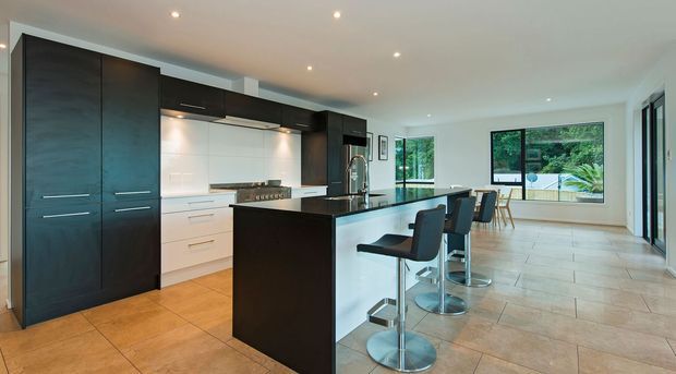 Contemporary open-plan kitchen with a large dark island, integrated dark cabinetry, white cabinetry and splashback, stainless-steel appliances, and three bar stools. The space opens toward a bright dining or living area with large windows and pale tiled flooring.