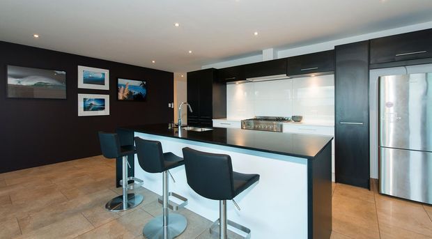 Contemporary open-plan kitchen with a large white island and integrated breakfast bar, black cabinetry, stainless-steel appliances, glossy white splashback, recessed ceiling lights, and dark feature walls. Four modern bar stools and framed artwork are visible, with warm-toned tiled flooring throughout.