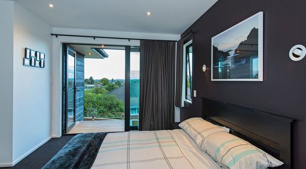A contemporary bedroom with a dark feature wall, upholstered bed, neutral checked bedding, dark carpet, recessed ceiling lights, and full-height glazed doors opening to a small balcony or outdoor area. The room overlooks a leafy suburban landscape and includes framed artwork and layered curtains.