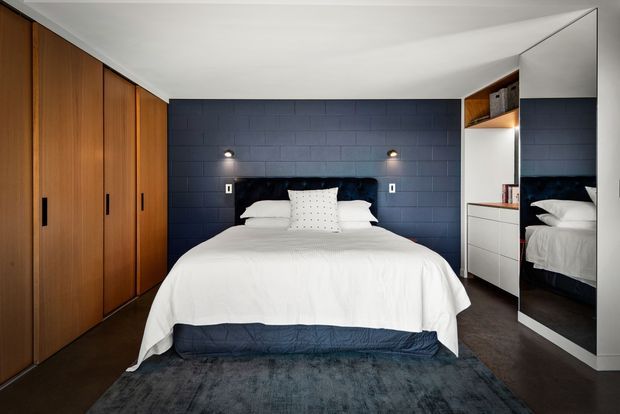 A compact contemporary bedroom with a neatly made double bed centred against a dark navy-blue tiled feature wall. Warm timber built-in wardrobes line the left wall, while white built-in drawers and open shelving sit beside a full-height mirrored panel on the right. Recessed and wall-mounted lighting, dark flooring, and a deep blue rug create a refined, moody contrast with the white bedding.