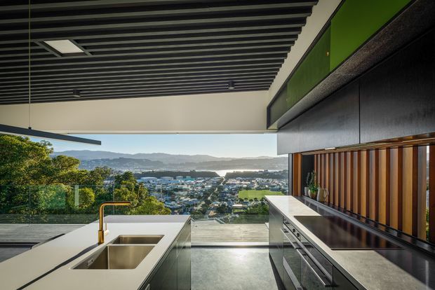 A contemporary kitchen with a large island, double stainless-steel sink and brass tap, integrated cabinetry, and a built-in cooking wall. Full-height glazing opens to an expansive elevated view over a suburban landscape and distant hills. Dark linear ceiling battens contrast with pale stone surfaces, while timber vertical slats and green upper cabinetry add warmth and colour.