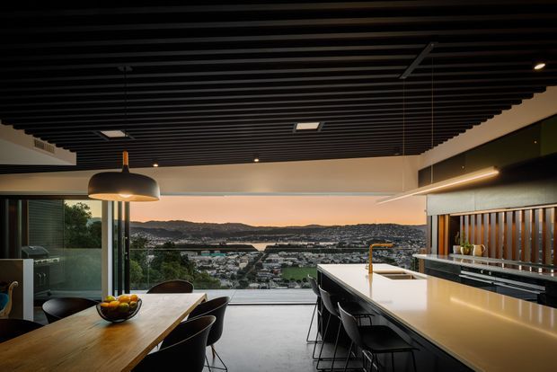 A sophisticated open-plan kitchen and dining area with expansive floor-to-ceiling glazing overlooking a coastal suburban landscape and hills at dusk. The space features a long pale stone island with integrated sink and brass-toned tap, dark cabinetry, black bar stools, a timber dining table, and a dramatic black slatted ceiling with contemporary linear lighting. The overall design is refined, modern, and strongly connected to the view.