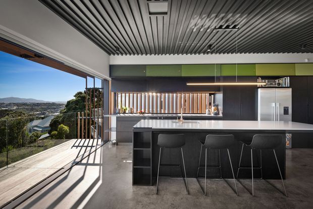 A contemporary open-plan kitchen viewed from an adjoining covered outdoor area or glazed opening. The kitchen features a large white island benchtop with three black stools, dark cabinetry, integrated stainless-steel appliances, a timber-toned vertical feature backsplash, and a linear pendant light. A striking slatted ceiling continues across the interior and exterior threshold, while full-height glazing opens to a timber deck with expansive suburban and hilly views.
