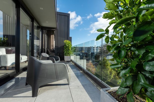 A contemporary apartment balcony or terrace with large-format grey paving, frameless glass balustrading, dark-framed floor-to-ceiling glazing, and sculptural outdoor lounge chairs. A leafy potted citrus tree occupies the foreground, with an urban neighbourhood visible beyond under a bright blue sky.