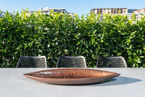 Outdoor dining setting with a long pale table, three dark woven-back chairs, and a decorative elongated copper-toned bowl in the foreground. A dense manicured hedge screens the setting, with modern multi-storey buildings visible beyond under a clear blue sky.