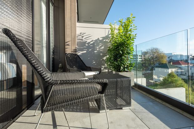 A sunny apartment balcony or terrace furnished with two dark woven lounge chairs and a matching square side table. The space has pale tiled flooring, dark-framed full-height glazing and sliding doors, a light rendered privacy wall, and a clear glass balustrade overlooking a suburban urban neighbourhood. A potted leafy tree adds greenery.