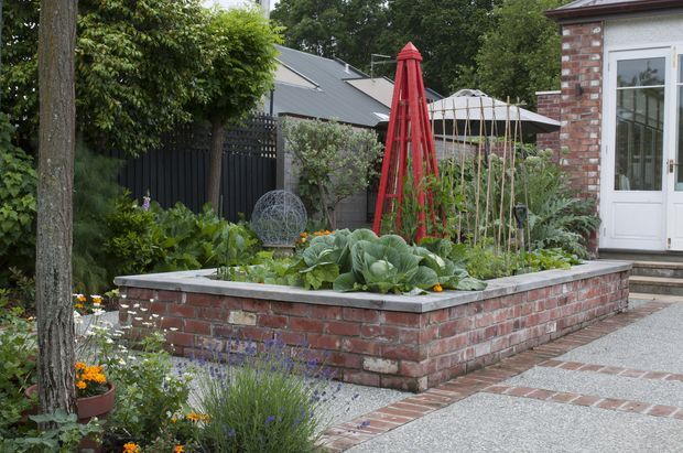 A landscaped courtyard garden with a raised brick vegetable planter bordered by paving. The garden includes leafy vegetables, trellises, a red garden obelisk, flowering plants, mature trees, and a brick outbuilding with white French doors. The setting has a refined traditional residential character.