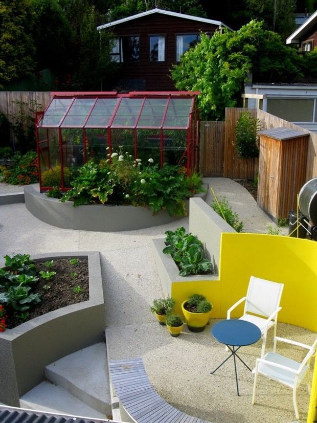 A landscaped urban courtyard garden with raised concrete planting beds, a red-framed glass greenhouse, paved paths, timber fencing, and a small outdoor seating area beside a bright yellow privacy wall. Dense greenery and a dark timber house are visible in the background.