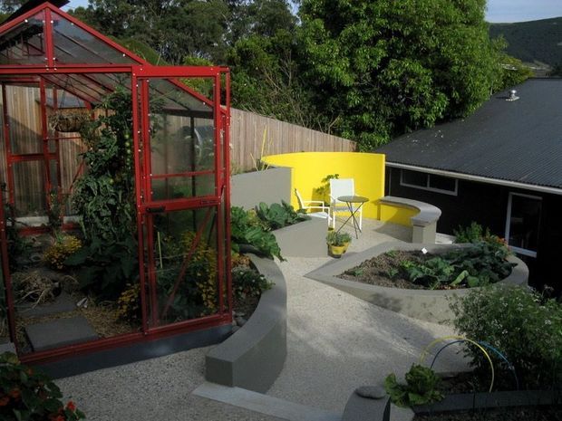 A landscaped courtyard garden beside a dark-roofed building, featuring a prominent red-framed glass greenhouse, curved raised concrete planting beds, pale textured paving, and a bright yellow curved garden wall with outdoor seating. Dense greenery and a sloping wooded landscape are visible beyond the property.