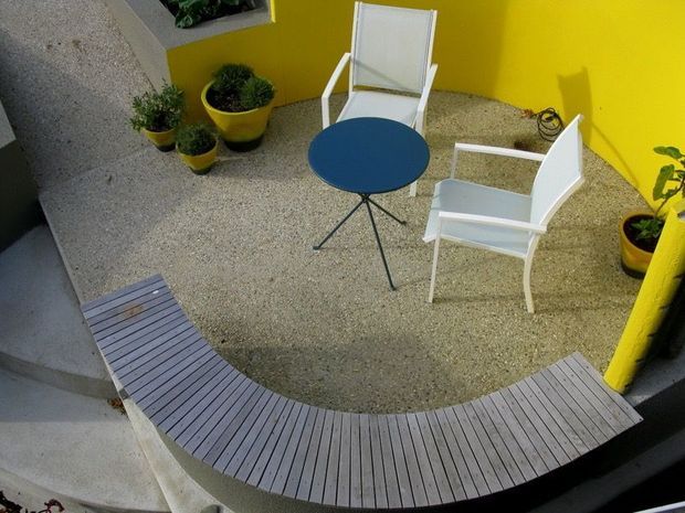 A compact outdoor courtyard or terrace with a curved slatted timber bench, two white outdoor armchairs, and a small round blue table. The space has bright yellow rendered walls, a speckled light aggregate floor, and several potted plants, creating a playful modern outdoor seating area.