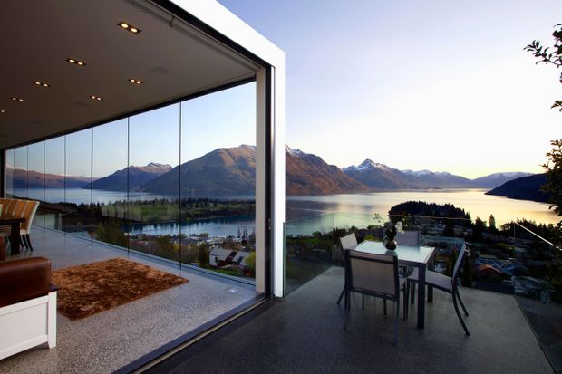 A modern open-plan living and dining space with floor-to-ceiling frameless glazing opening onto an outdoor terrace. The interior overlooks a dramatic alpine lake and mountain landscape, with a dining table and chairs on the terrace, a partially visible sofa and rug indoors, recessed ceiling lighting, and a crisp white structural frame.