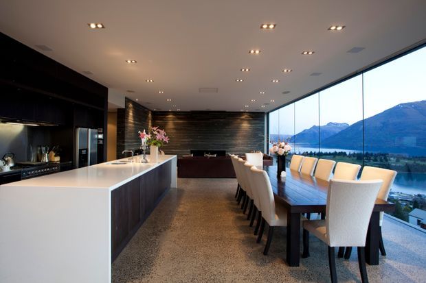 A spacious contemporary open-plan kitchen and dining area with a large white island, dark timber cabinetry and feature wall, polished concrete flooring, and a long dining table with upholstered chairs. Full-height glazing opens to an expansive mountain and lakeside view. Recessed ceiling lighting and restrained detailing create a refined, luxurious interior.
