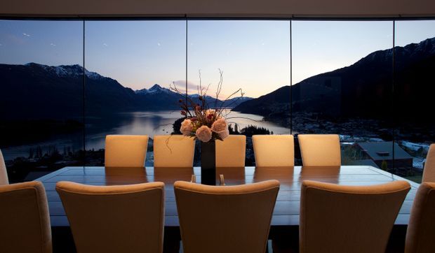 A formal dining area positioned in front of expansive floor-to-ceiling glazing, with a long rectangular dining table, upholstered tan dining chairs, and a central floral arrangement. The glazing frames a dramatic mountain and lake landscape at dusk, creating a refined, scenic interior setting.
