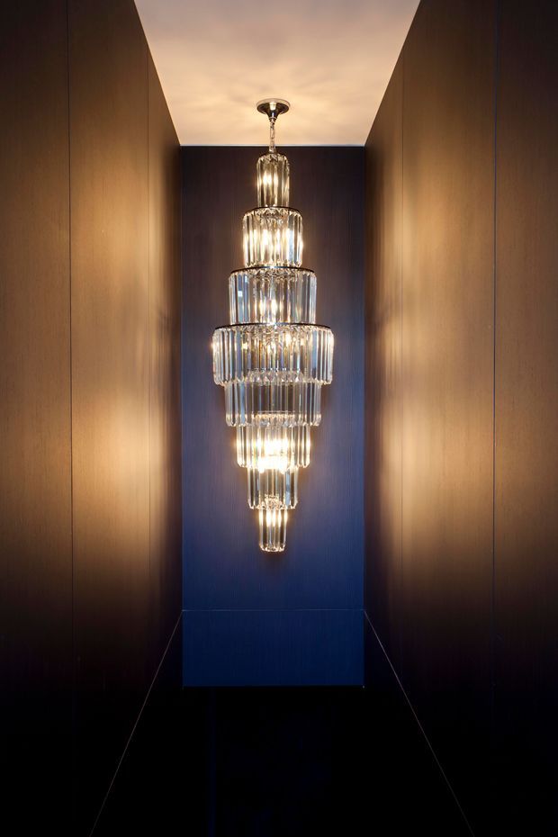 A narrow, dramatic interior alcove or hallway featuring a large tiered crystal pendant chandelier suspended against a deep navy-blue feature wall. Warm wood-toned wall panels line both sides, while the dark glossy floor and focused lighting create a luxurious, high-contrast atmosphere.