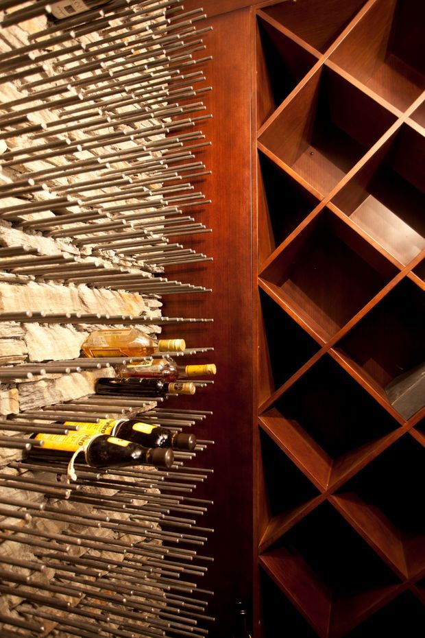 A dramatic wine cellar interior featuring extensive dark timber wine racking with diamond-shaped compartments on the right and a sculptural wall-mounted bottle storage system using numerous horizontal metal rods on the left. Several wine bottles are displayed among rough stone-textured surfaces, creating a dark, warm, luxurious atmosphere.