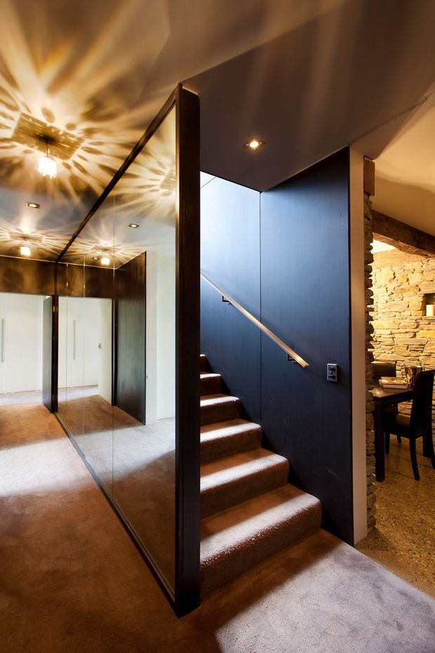 Interior view centred on a compact carpeted staircase with a dark blue wall and timber handrail. A full-height mirrored wall reflects the hallway and decorative ceiling light, while an opening to the right reveals a dining area with a rough stacked-stone feature wall and dark furnishings. The space combines dramatic warm lighting, dark finishes, and textured natural stone.