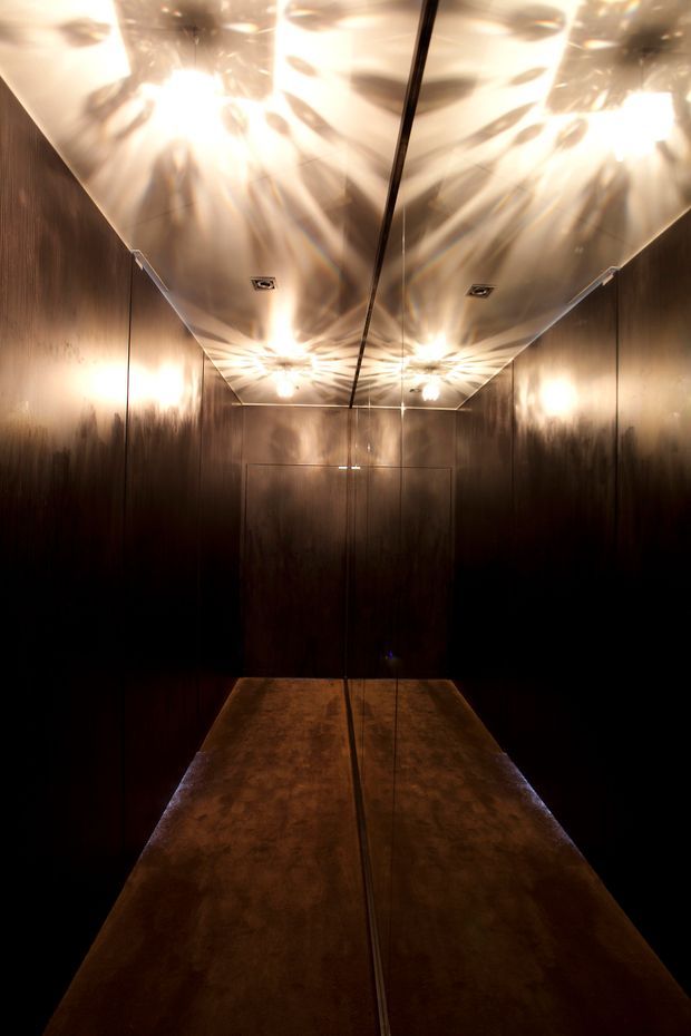 A narrow, dark lift or enclosed hallway lined with dark reflective wall panels and a mirrored ceiling. Warm decorative lighting creates dramatic radial reflections across the ceiling and walls, while the floor appears to have a mottled bronze or dark stone finish. The space has a luxurious, moody, highly reflective character.