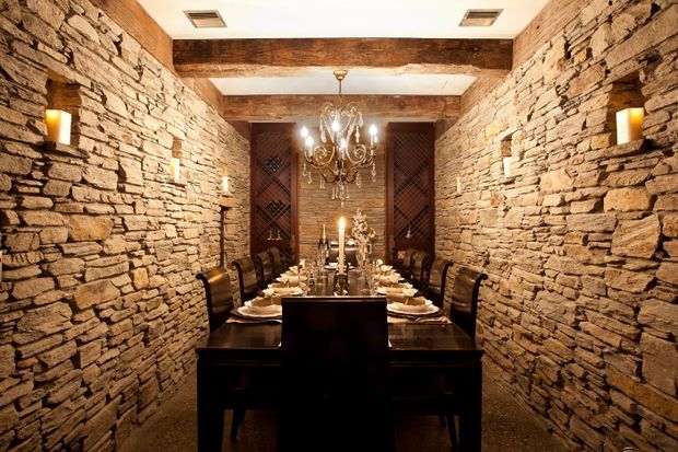 A dramatic, intimate dining room with a long dark timber dining table and matching chairs, set for a formal dinner. Rough stacked-stone walls run along both sides, with exposed dark timber ceiling beams, warm wall sconces, a large ornate chandelier, and candlelight creating a rustic yet refined atmosphere.