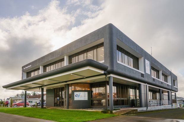 Exterior view of a two-storey commercial building with a rounded-corner dark grey concrete or rendered facade, extensive horizontal glazing, and a prominent projecting flat canopy supported by slim columns. The building sits on a landscaped site with a paved entrance and parking visible around it.
