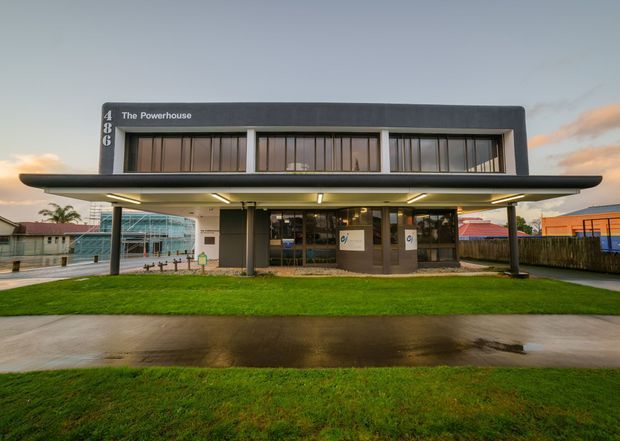 Exterior view of a contemporary two-storey commercial building named The Powerhouse. The building has a dark rectangular upper volume with extensive vertical glazing, a broad projecting flat canopy supported by slender columns, and a glazed ground-floor entrance. It sits behind a wet paved forecourt and lawn in an urban or suburban setting at dusk.