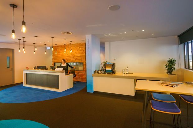 A contemporary commercial reception and waiting area with a white reception desk, long service counter, coffee station, and small meeting table. The space features a warm exposed brick feature wall, blue and dark carpet zones, pendant lighting, white ceilings, and blue upholstered stools. Large windows provide daylight along the right side.