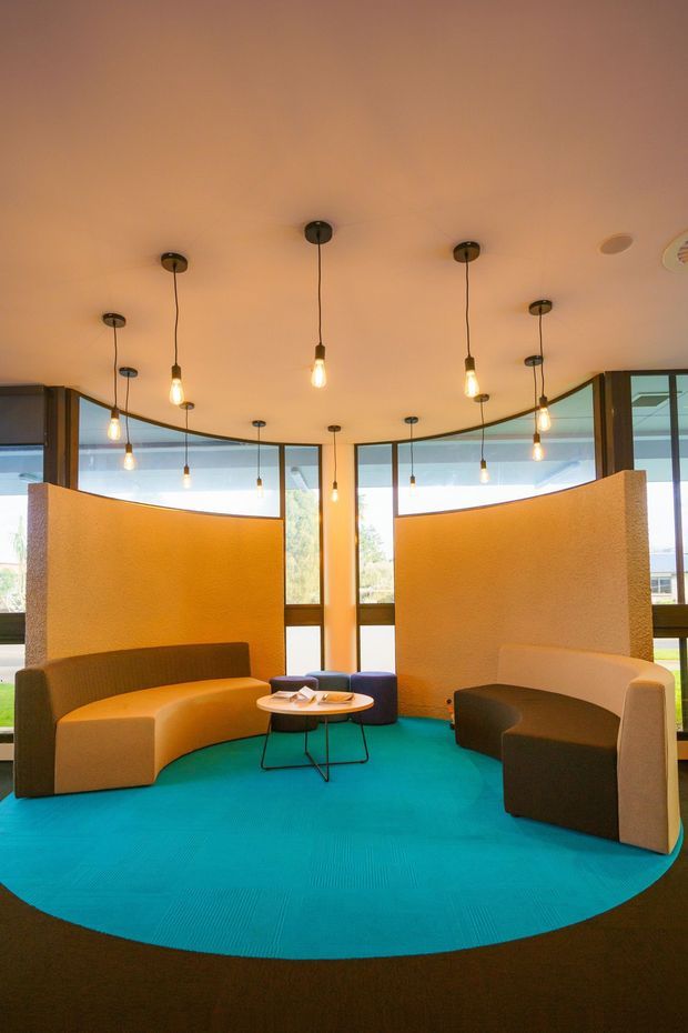 A bright, curved seating nook with sculptural upholstered banquettes arranged around a small round coffee table. The space features a vivid turquoise circular carpet, warm beige textured partition walls, extensive floor-to-ceiling glazing, and numerous suspended exposed-bulb pendant lights. The composition suggests a contemporary commercial lounge or reception area.