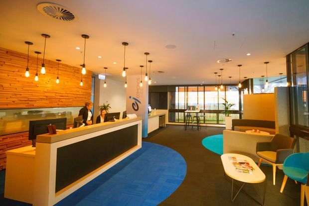 A contemporary commercial reception and lounge area with a long angular reception desk, illuminated pendant lights, a warm horizontal timber feature wall, carpeted flooring with blue and turquoise circular accents, and seating arranged beside expansive glazed windows.