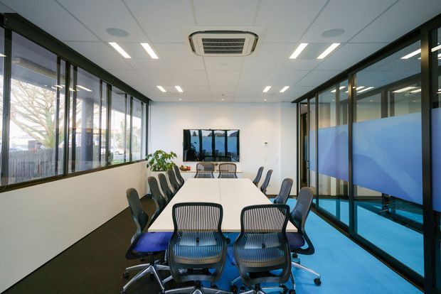 A bright contemporary office meeting room with a long white conference table surrounded by ergonomic office chairs. Full-height internal and external glazing lines both sides, with blue translucent privacy graphics on the right. The room has white walls, a suspended acoustic tile ceiling with recessed linear lighting and a ceiling cassette air-conditioning unit, plus dark flooring and blue carpet or vinyl flooring. A wall-mounted display is visible at the far end.