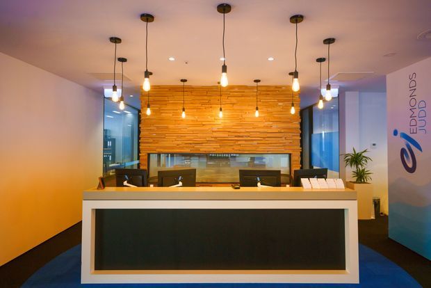 A contemporary commercial reception area featuring a large white-framed reception desk with a dark front panel, multiple computer workstations, and a warm horizontally stacked timber feature wall behind the counter. Numerous exposed pendant bulbs provide warm ambient lighting, while glass partitions and a branded wall panel reinforce the office or corporate setting.
