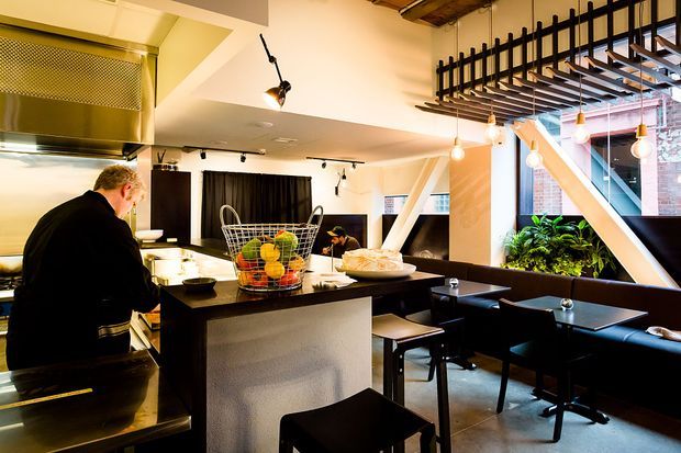 A stylish open-plan restaurant interior combining a commercial kitchen, central service island, dining tables, banquette seating, and a planted area beside large windows. The space features exposed timber ceiling structure, a black metal screen and pergola-like overhead grid, warm pendant bulbs, dark furniture, stainless-steel kitchen surfaces, and bold white structural beams. The atmosphere is warm, urban, and industrial-leaning.