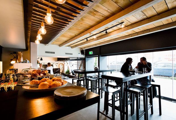A contemporary café or restaurant interior with a long bar and food display in the foreground, high communal tables and stools beside full-height glazing, and a partially visible kitchen or service area at the rear. The space features exposed timber ceiling beams and boards, black-painted structural and joinery elements, track lighting, pendant bulbs, and a warm industrial hospitality atmosphere.