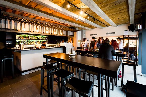 A warmly lit contemporary bar or hospitality venue with a long dark timber communal table and black stools in the foreground. The space features a backlit liquor display and bar counter, exposed timber ceiling boards, substantial pale timber beams, and a striking slatted timber ceiling feature above the bar. Patrons gather around the rear counter, while large glazed openings and dark-framed detailing add an urban industrial character.