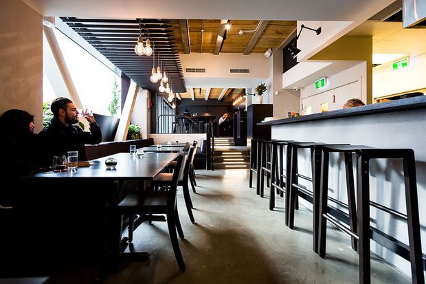 A contemporary restaurant or café interior with a long communal bar and black metal stools on the right, dining tables and chairs on the left, and a raised rear area reached by steps. The space features polished concrete flooring, dark timber ceiling slats and beams, pendant globe lighting, black-framed glazing, and a warm neutral palette with strong linear detailing.