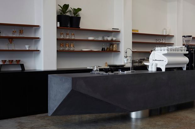 A sophisticated commercial-style coffee bar with a large dark sculptural island counter, integrated sink and tap, white espresso machine, and open dark timber shelves displaying cups, glasses, and coffee equipment. The space has white plaster walls, black cabinetry, concrete-look finishes, and potted plants, creating a refined contemporary café atmosphere.