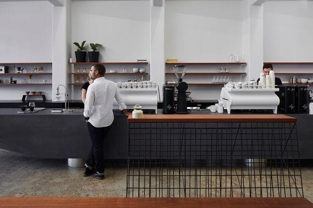A contemporary café interior viewed from the customer seating area, with a long black service counter, espresso machines, grinders, open timber display shelves, and a central island clad in a black metal grid. White walls, concrete flooring, timber accents, and potted plants create a clean, understated coffee-shop setting.