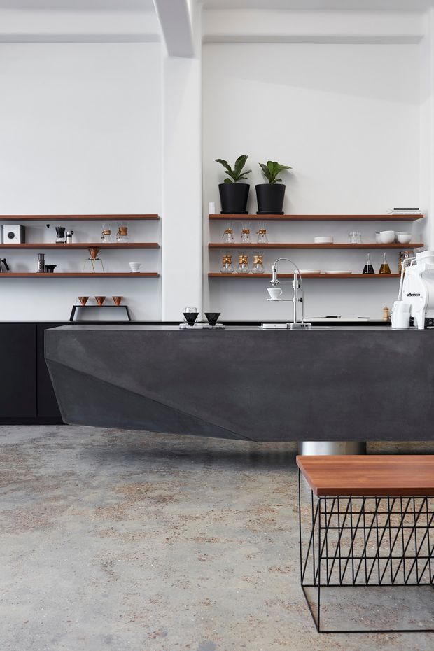 A minimalist contemporary kitchen with a dramatic charcoal island featuring an angular sculptural base, black cabinetry, dark timber open shelving, and white walls with exposed structural beams. The island includes a stainless-steel sink and tall curved tap, while the shelves display glassware, ceramics, small appliances, and potted plants. The space has a raw, mottled concrete floor and a timber-topped wire-frame coffee table in the foreground.