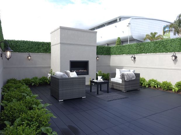 A refined rooftop or elevated outdoor entertaining terrace enclosed by rendered privacy walls and clipped hedging. The space features dark decking, a built-in outdoor fireplace, wicker lounge seating with pale cushions, a low coffee table, wall-mounted lanterns, and landscaped planting. A contemporary building is visible beyond the enclosure.