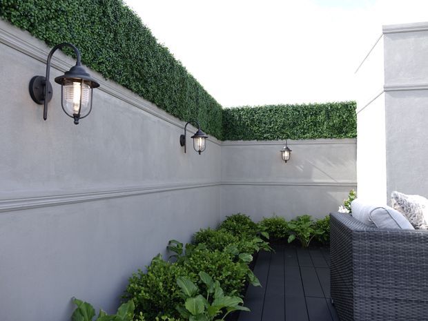 A narrow enclosed outdoor terrace or courtyard with pale rendered walls, decorative horizontal moulding, dense green hedge screening along the wall tops, black wall-mounted lanterns, dark decking, and lush planted garden beds. A woven outdoor sofa occupies the right side.