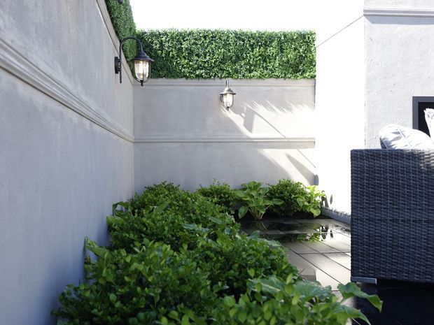 A compact enclosed courtyard or terrace with pale rendered walls, decorative black wall lanterns, dense green hedge planting, and dark outdoor wicker furniture at the right edge. Sunlight creates strong shadows across the walls and paving, giving the space a private, leafy outdoor character.