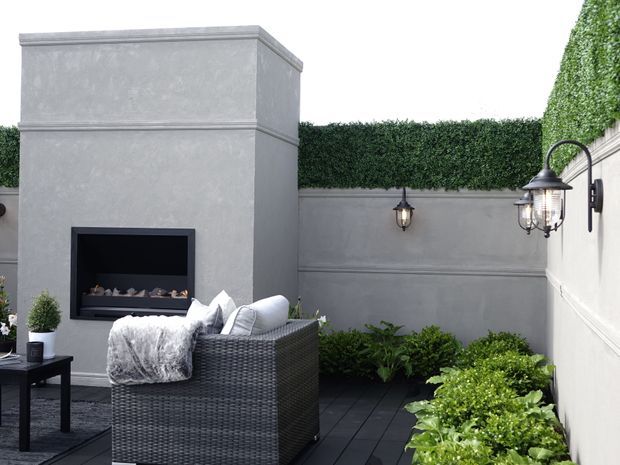 A refined enclosed outdoor courtyard or terrace with pale rendered walls, dark tiled flooring, clipped hedge screening, and built-in raised planting beds. A large square outdoor fireplace forms the focal point, complemented by traditional wall lanterns and dark woven outdoor seating with light throws and cushions.