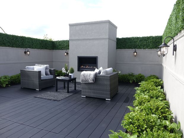 A refined rooftop or enclosed courtyard outdoor lounge with dark decking, two woven armchairs, a small side table, wall-mounted lanterns, clipped hedge screening, raised planting beds, and a built-in outdoor fireplace set into a rendered feature wall.