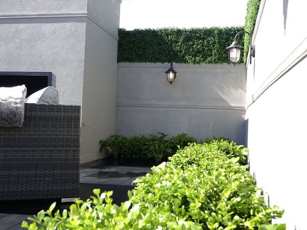 A bright, enclosed courtyard or outdoor terrace with rendered grey walls, dark tiled paving, clipped hedge screening, low planted borders, and traditional wall-mounted lanterns. A dark woven outdoor lounge chair is partially visible at left, creating a private, manicured garden setting.