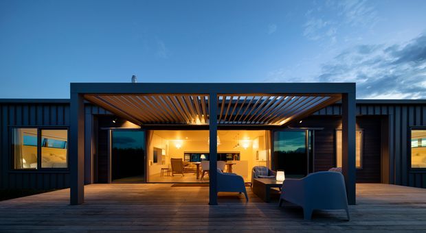 Contemporary single-storey dark-clad home viewed from a broad timber deck at dusk. A deep covered outdoor living area with a timber slatted pergola frames a warmly lit interior containing a kitchen and dining area, while dark-framed glazing and integrated outdoor seating create a strong indoor–outdoor connection.