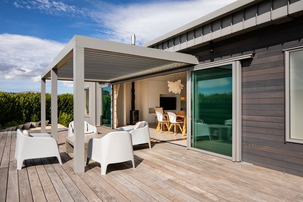 A contemporary outdoor terrace with weathered timber decking, white lounge chairs, and a covered pergola extending from a modern home. The open sliding glass doors reveal a bright dining and living area, while dark horizontal timber cladding contrasts with the pale structural framing and expansive glazing.