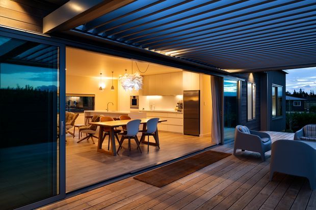 A contemporary open-plan kitchen and dining area opens directly onto a timber deck through expansive sliding glass doors. The warmly lit interior features pale timber cabinetry, a central island, dining table, pendant lights, and stainless-steel appliances. The covered outdoor deck has a slatted timber ceiling, lounge seating, and dark vertical exterior cladding, creating a strong indoor-outdoor connection at dusk.