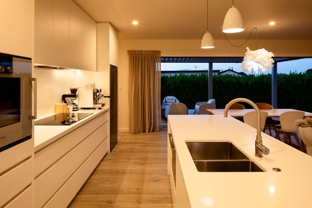 Warm contemporary open-plan kitchen with sleek white handleless cabinetry, integrated appliances, a large white island with double stainless-steel sink, and pendant lighting. The kitchen opens to a dining and outdoor entertaining area through wide glazing, with timber flooring and neutral furnishings.