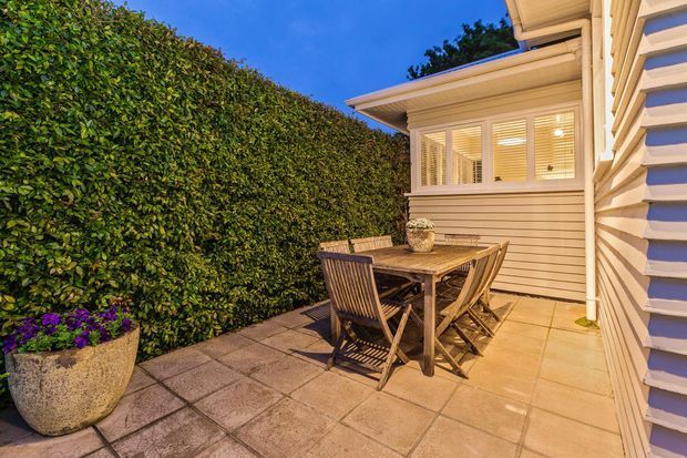 A compact outdoor dining patio beside a light-coloured weatherboard house. The paved terrace contains a wooden dining table and chairs, with a tall manicured hedge screening one side, a large planter, and an enclosed glazed room with horizontal blinds in the background. The scene is warmly illuminated at dusk.